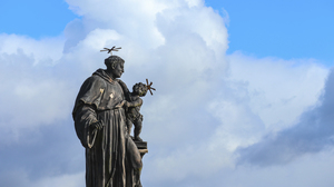 Dark sculpture of Anthony of Padua holding a small child with a blue sky background.