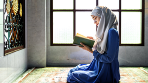 Muslim woman in a blue dress and a grey hijab sitting on a colorful carpet in front of a big window inside a mosque, reading from the Koran.