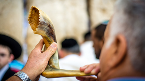 An elderly man holding a ram's horn (shofar) to his lips, with other people in Jewish attire in the background.