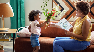 Woman sitting on a couch reading a card while a kid gives her a bouquet of flowers.