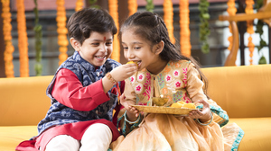 Two kids in festive clothing. Kid on left is in blue and red kurta and is feeding sweets to child on right. Child on right is wearing pink festive clothing.