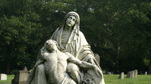Old, stone statue of Mary holding Jesus in her arms in a cemetery with tall, green trees in the background.