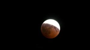 A partially eclipsed Moon in the night sky. A little over half of the Moon is covered by a reddish shade.