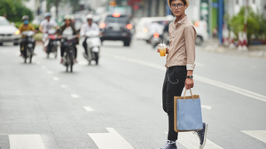 Young man wearing a stylish hat and glasses and carrying shopping bags crossing a city street.