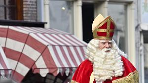 Sinterklass with a white beard and red robes and a red crown arriving on horseback. Blurred store fronts and building in the background.