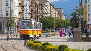 A yellow tramway is winding its way through the aging cityblocks of Sofia, Bulgaria. The street shows blue mountains in the horizon.