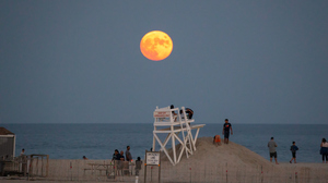 A large glowing yellow Full Moon rising above the ocean and lifeguard towers and people watching on Long Beach, New York.