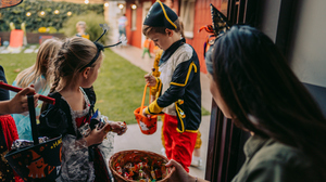 Children in costumes trick-or-treating at a house during Halloween. A woman holds a basket of candy for children who are carrying their own Halloween-themed buckets.