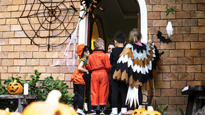 Children in Halloween costumes trick-or-treating at the door of a decorated house with pumpkins and spider webs.