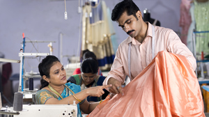 Two garment workers, one sitting down and wearing a blue sari and the other, standing and wearing a peach shirt discussing over an orange fabric.