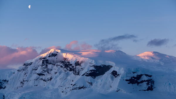 Snow-capped mountains in sunset with a Half Moon above them in a blue sky.