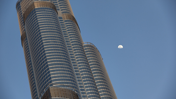 Close-up of a skyscraper with a gibbous Moon next to it in a blue sky. The top half of the Moon is illuminated.