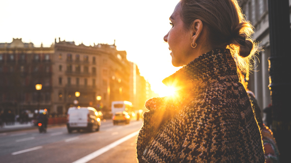 A woman looks out onto a city street with the setting Sun illuminating her from behind.