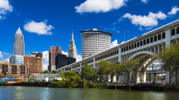Downtown Cleveland skyline with the Detroit Superior Bridge and Cuyahoga River in the foreground, and a deep blue sky with puffy cumulus clouds in the background.