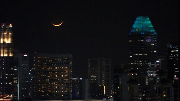 An orange Waxing Crescent Moon setting over Singapore downtown. The millennia tower top is illuminated with blue lights. Other towers and buildings of financial district are illuminated by lights at night.