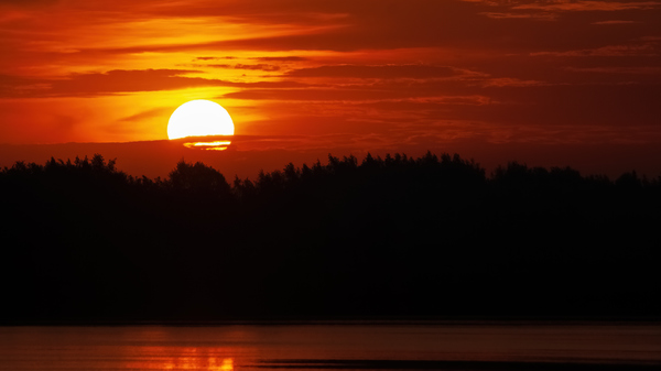 Bright orange Sun setting behind dark silhouette of forest and river.