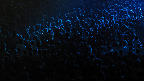 Crowd of people standing in near-darkness, all looking in one direction, the contours of hundreds of heads illuminated by a faint blue light..