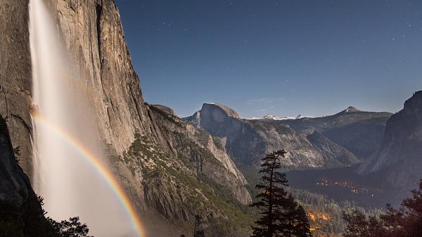 Image of a waterfall at night with one bright moon bow and a faint moon bow above it. Dark skies and city lights in the background.