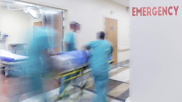 A team of doctors and nurses hurriedly pushing bed with a patient down a hospital corridor, with the word emergency written on the wall in large red letters.