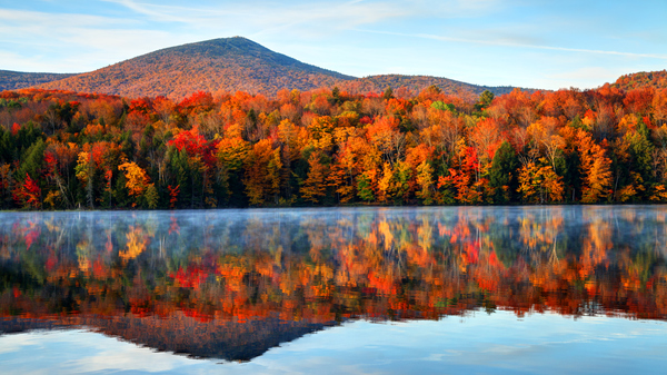 Image of colorful fall foliage reflecting in a lake with blue sky and a hill in the background.