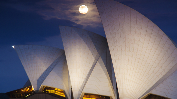 A bright white Full Moon rising behind the white curved sails of the Sydney Opera House in Sydney, Australia.