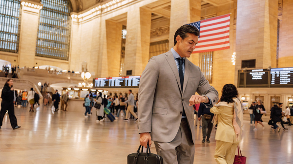 Business man in a suit glancing at his watch as he walks through a busy train station with more people and an American flag in the background.