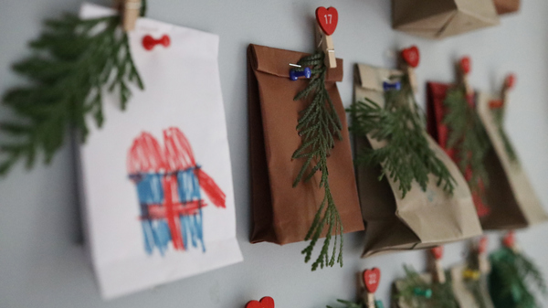 Close-up of a handmade advent calendar with small bags, greenery, and numbered heart-shaped clips on a wall. 