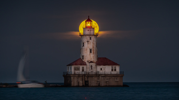 A beautiful night shot of the harvest full moon as it aligns with the center top of the abandoned historic light house tower along Lake Michigan in Chicago as sailboat passes by on the dark water.