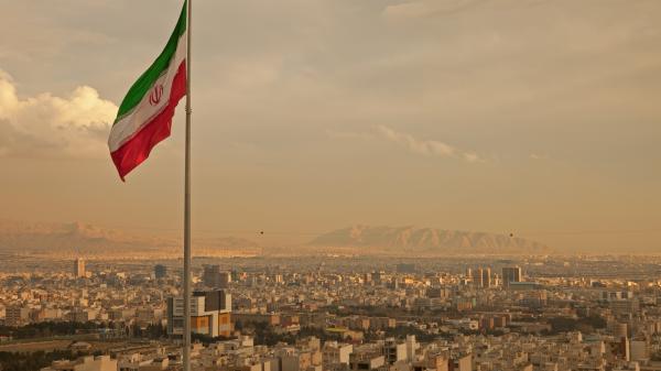 Iran flag waving in the wind above the skyline of Tehran lit by the orange glow of sunset.