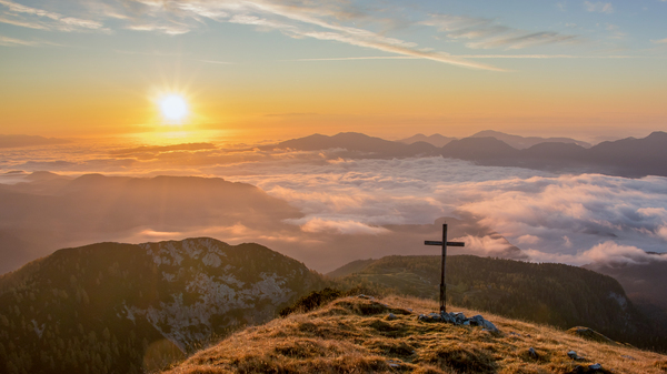Landscape view of grassy mountain tops peaking above the clouds during sunset, the mountain top in front with a simple cross perched on top.
