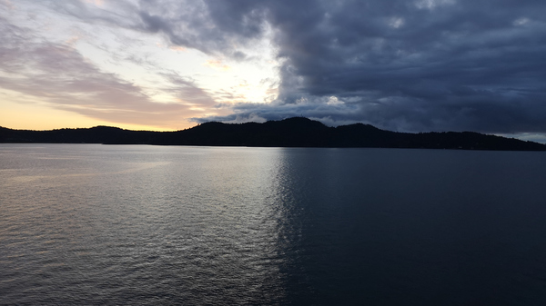 Body of water with hills in the background at twilight, with a clear sky on the left and a dark, cloudy sky on the right.