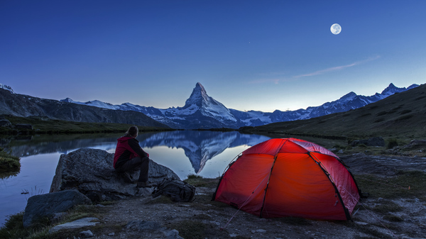 An illuminated tent under Full Moon at Matterhorn in Switzerland. A man is sitting by a lake, and the mountain is reflected in it.