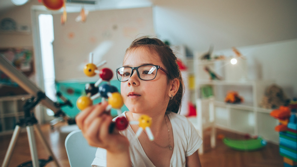 A young girl wearing glasses examines a molecular model in a cozy learning space with toys and a telescope.