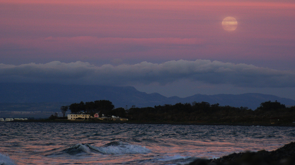 A yellow Full Moon rising ver some houses lit up by the setting Sun with the waves lapping.