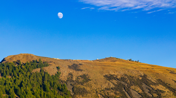Side view of a sparsely vegetated mountain with some buildings on top and a Gibbous Moon above it in the blue daytime sky.
