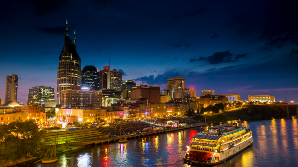 Skyline von Nashville, Tennessee, USA in der Abenddämmerung, mit hell beleuchteten Gebäuden und einem Flusschiff im Vordergrund.