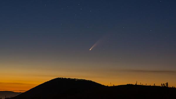 Comet in the dark sky over a mountain with a setting/rising Sun in the background.