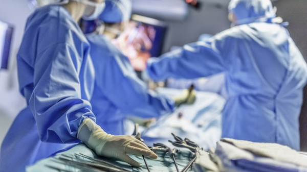 Surgical nurse reaching for an operating tool in the foreground while surgeons operate on a patient in the background.