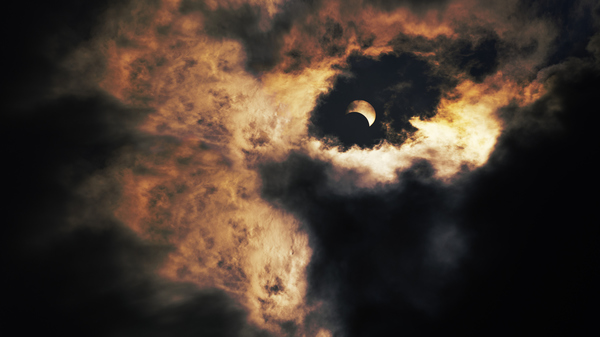 Image of the sky with lots of dark and orange clouds. In the middle of the clouds is the Sun partially covered by the Moon.