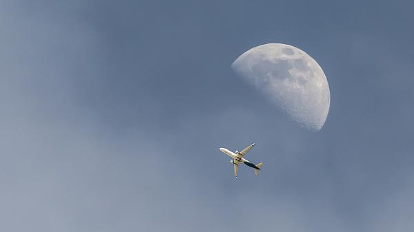 Underside of an airplane visible in front of a half Moon during the day with a blue sky in the background.