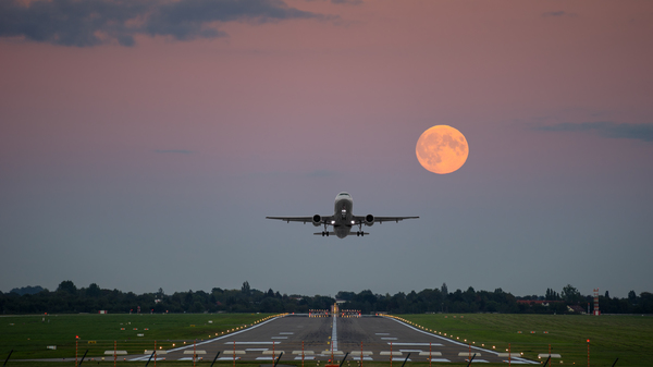 The Full Moon rising in the sky over a lit up runway with yellow lights and a plane taking off straight ahead.