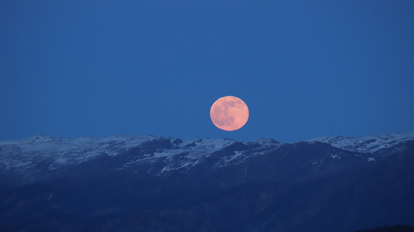 A Full Moon rises over snowy mountains.