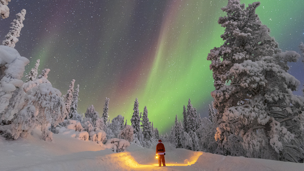 A person, Santa, in a red suit holding a lantern stands on a snowy path under a vibrant aurora borealis in a winter forest.