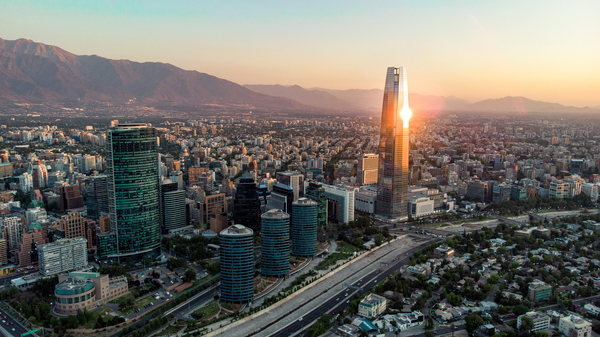 Cityscape of Santiago, Chile at sunset with the Andes mountains layering behind the high rise buildings. The buildings are standing close and a big tower reflecting the Sun is in the right, top corner of the image. The sky is pastel yellow and blue.