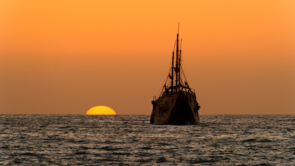 Untergehende Sonne mit Meer und Segelschiff im Vordergrund.
