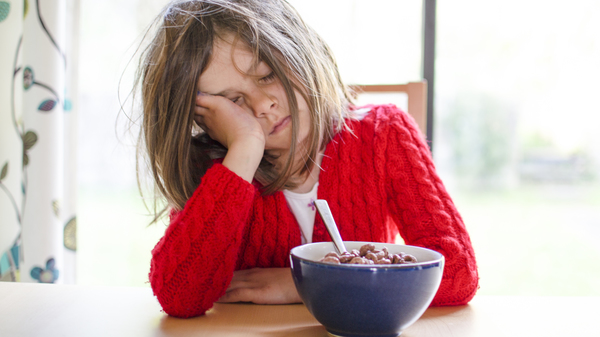 A child in a red jumper leaning their head in their hand and trying to eat a bowl of cereal.