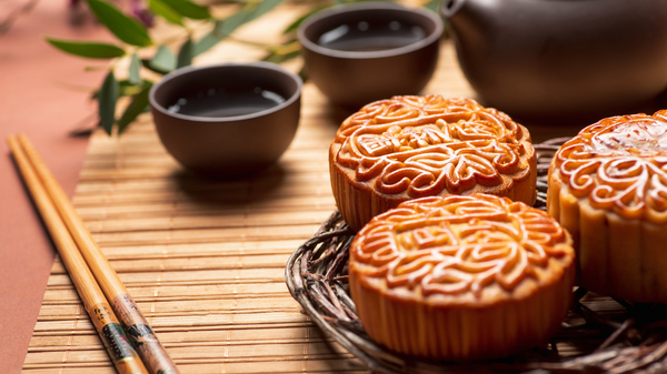 An image of three mooncakes, traditional Chinese pastries served during the Mid-Autumn Festival, alongside a teapot, two teacups, and a pair of chopsticks.