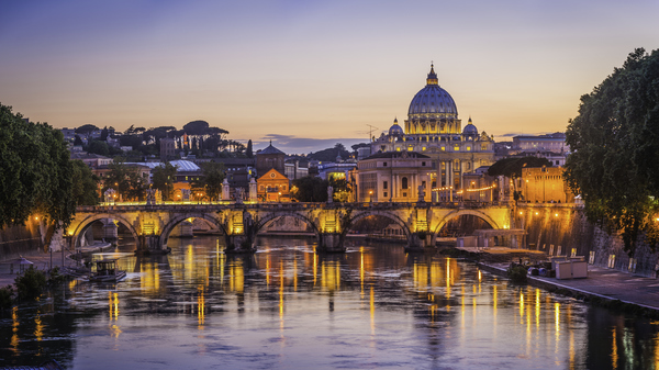 Vatican City at sunset, St. Peter's Basilica reflecting in the Tiber River with illuminated bridge foreground.