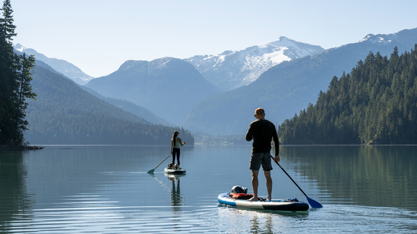 Two people standing on SUPs on a beautiful lake surrounded by wooded mountains.