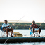 Two smiling men sitting in camping chairs by a river and holding fishing rods.